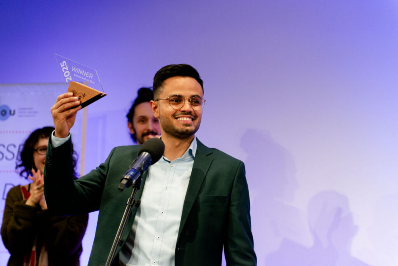 HOOU Science and Art Slam 2026 Der strahlende Sieger des Slams von 2025 hält seinen Pokal mit dem rechten Arm in die Höhe.